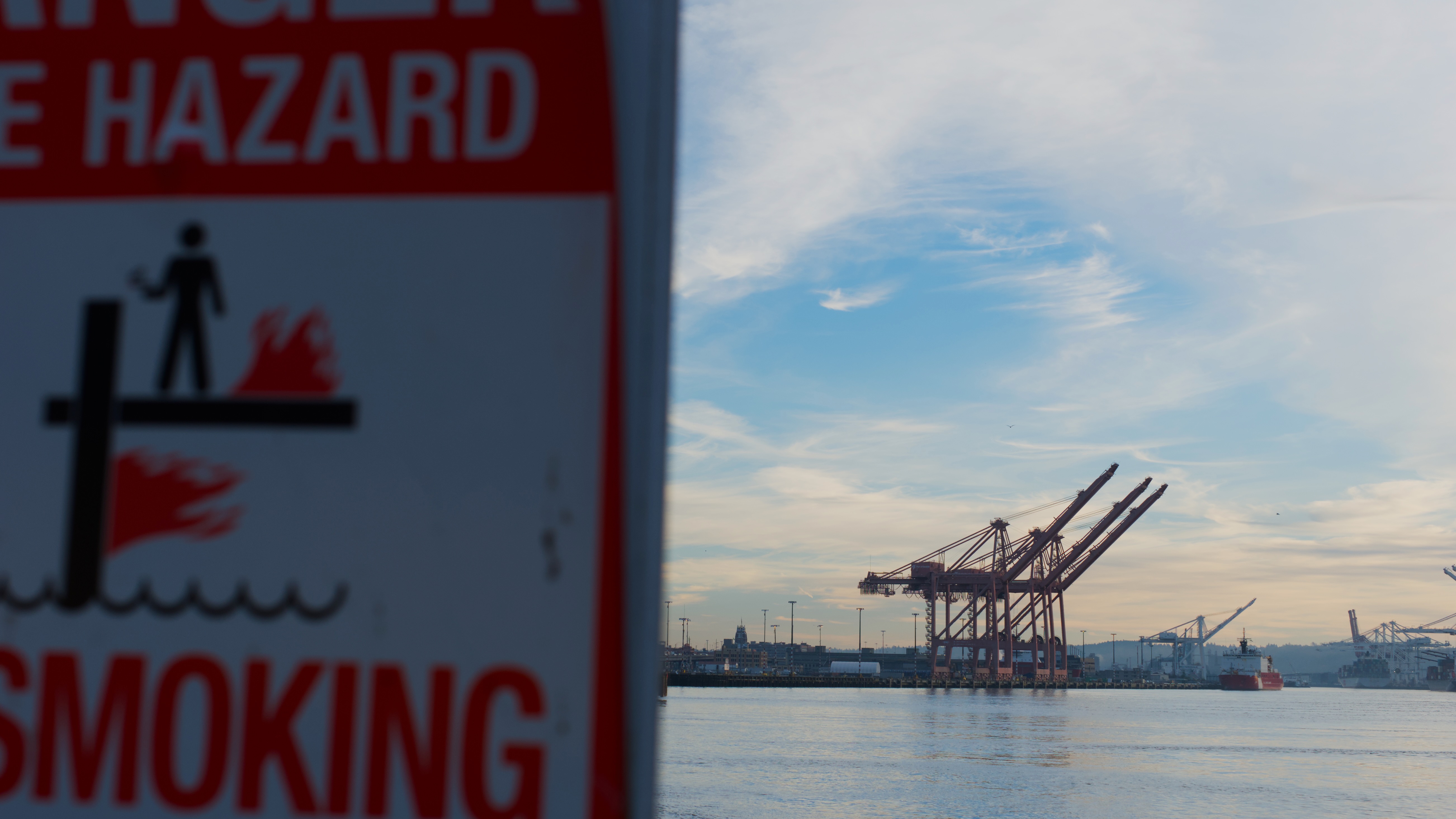 A fire hazard warning sign in the foreground with shipping container cranes visible across the water under a cloudy sky at dusk.