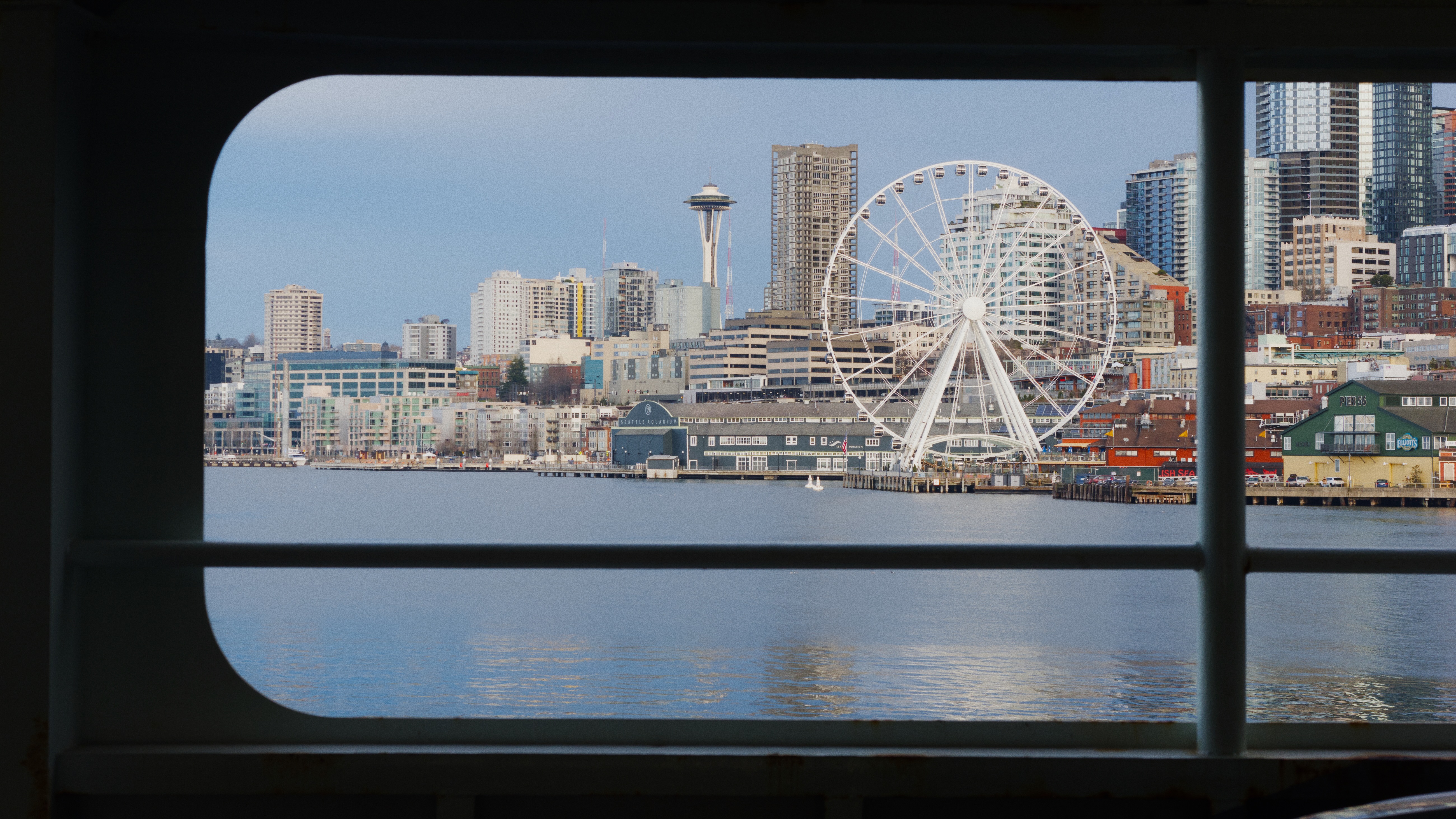 Seattle waterfront skyline featuring the Space Needle and Great Wheel ferris wheel, viewed through a ferry window across Elliott Bay.