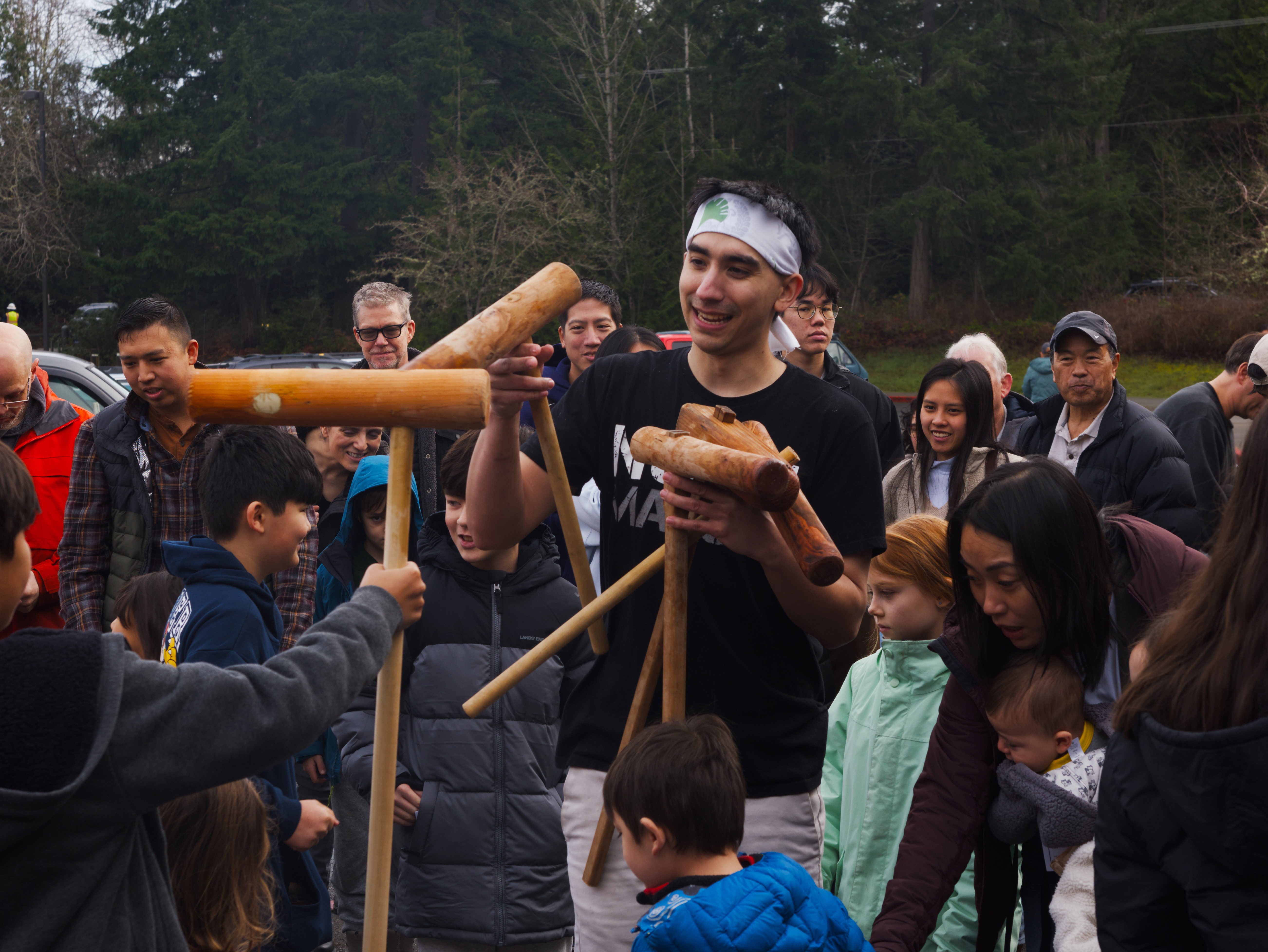 A group of people gathered outdoors watching a young man wearing a headband demonstrate mochi pounding with large wooden mallets (kine) at what appears to be a traditional mochitsuki event.